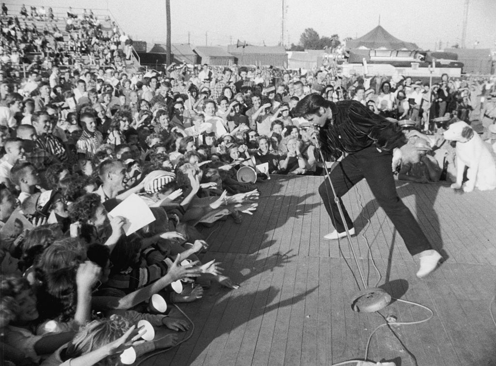 circa 1957: American singer and actor Elvis Presley (1935-1977) performing outdoors on a small stage to the adulation of a young crowd. (Photo by Hulton Archive/Getty Images)